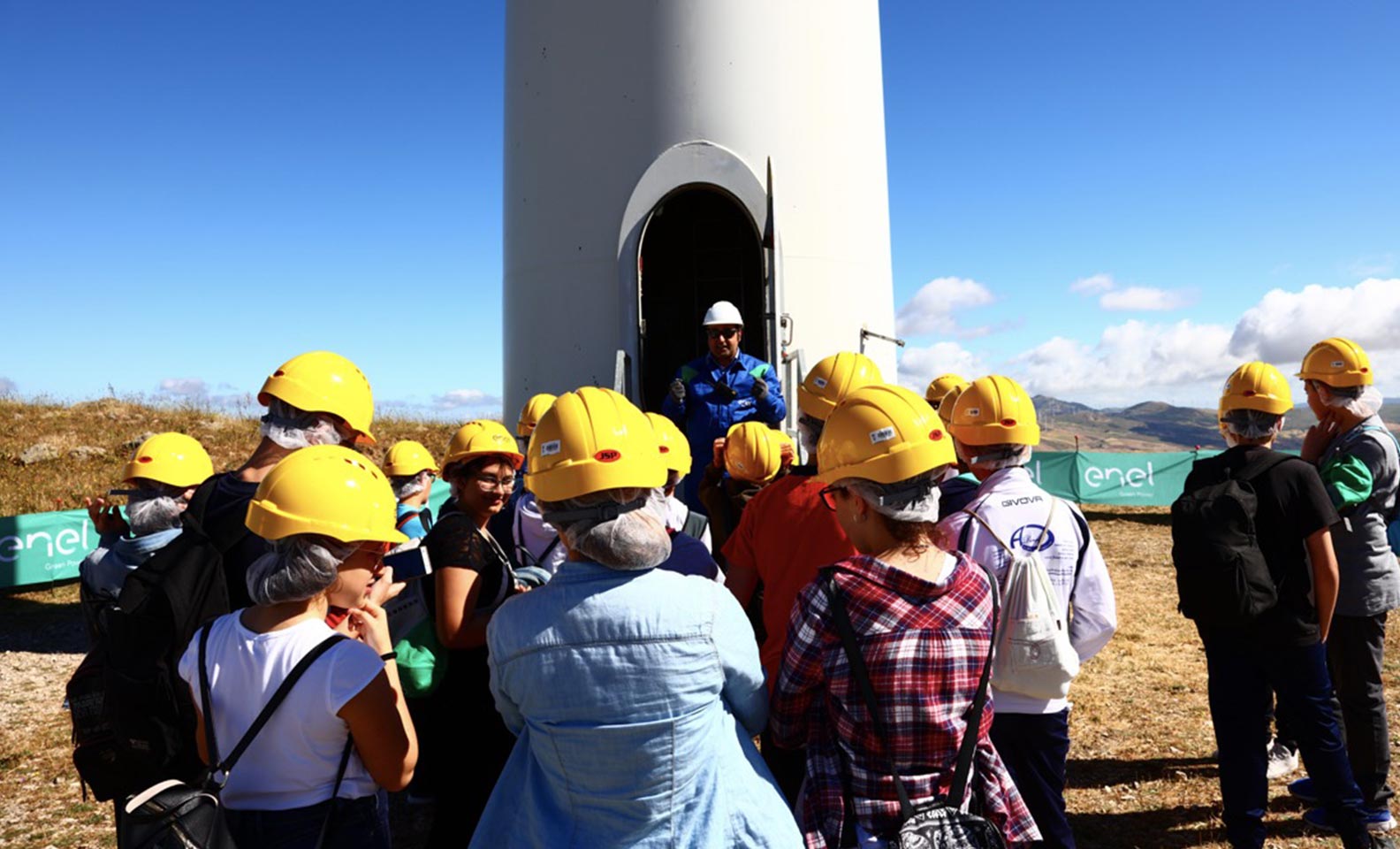 Homens e mulheres com capacete amarelo em frente de uma lâmina de turbina eólica